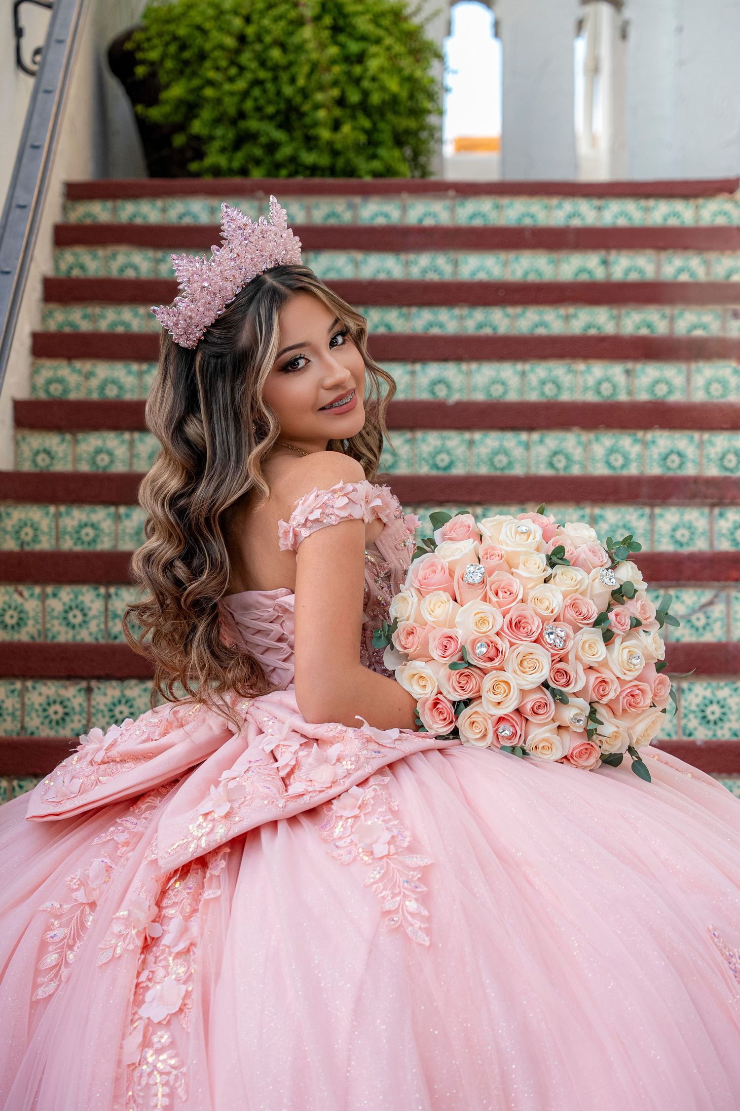Young woman in pink quinceañera ball gown with floral crown holds rose bouquet on decorative tiled stairs.