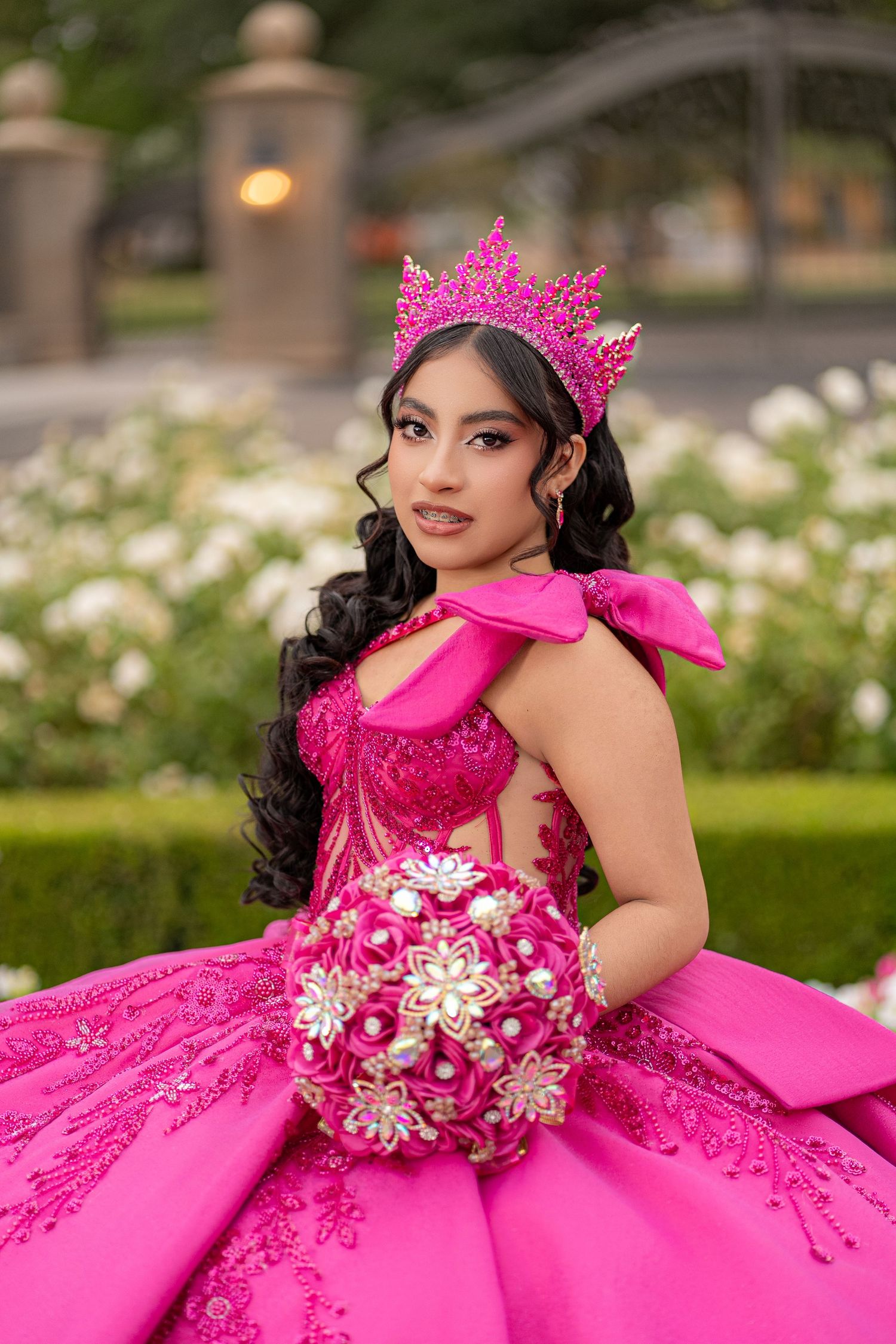 Young woman in hot pink quinceañera gown and crown holds floral bouquet, posing in garden.