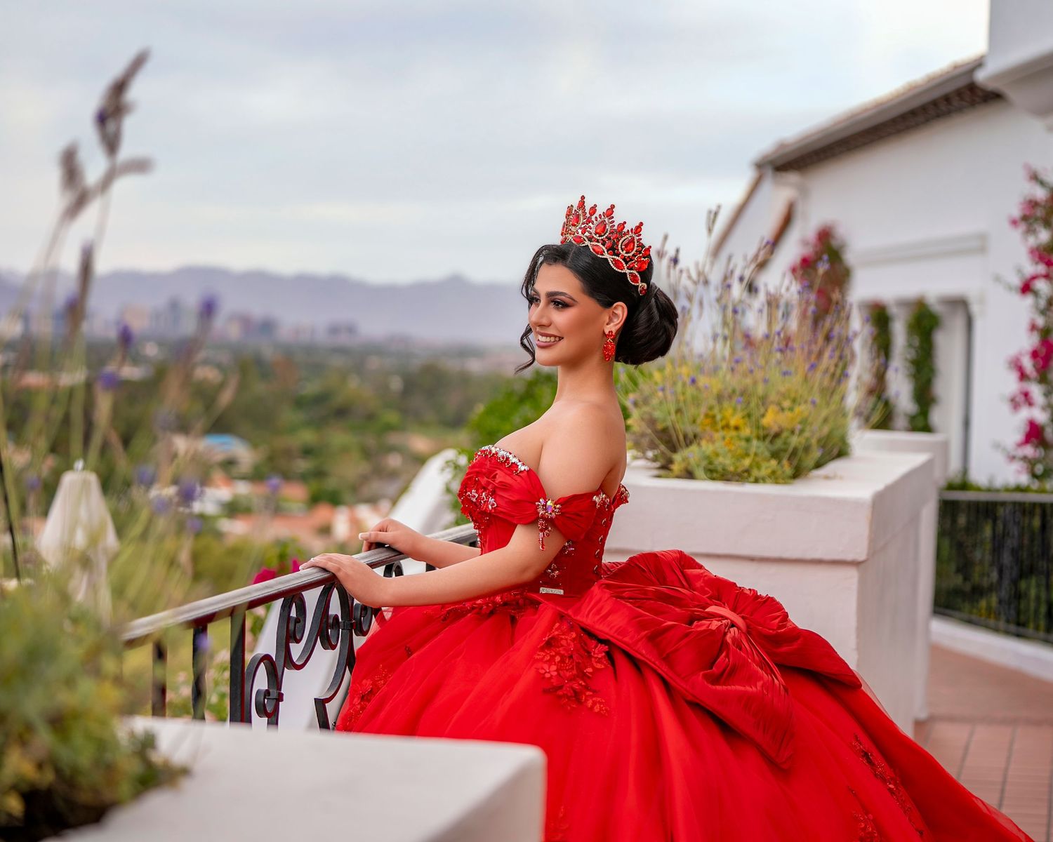 Young woman in red off-shoulder quinceañera gown with crown poses on balcony with mountain views.