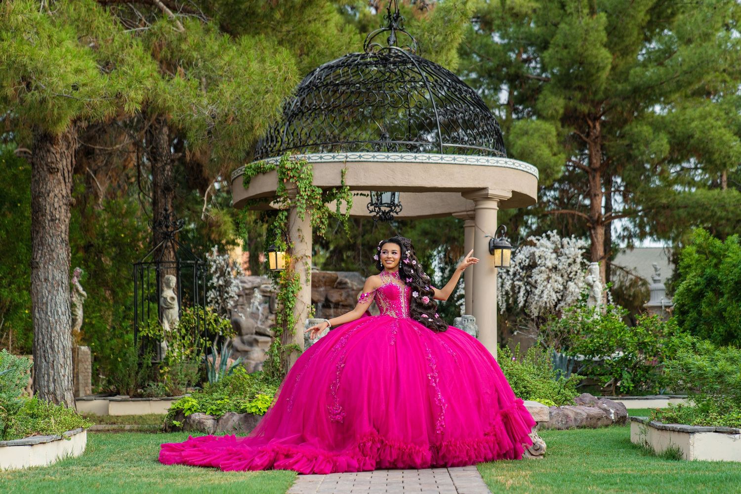 Young woman in hot pink quinceañera ball gown poses arms wide under decorative iron gazebo.