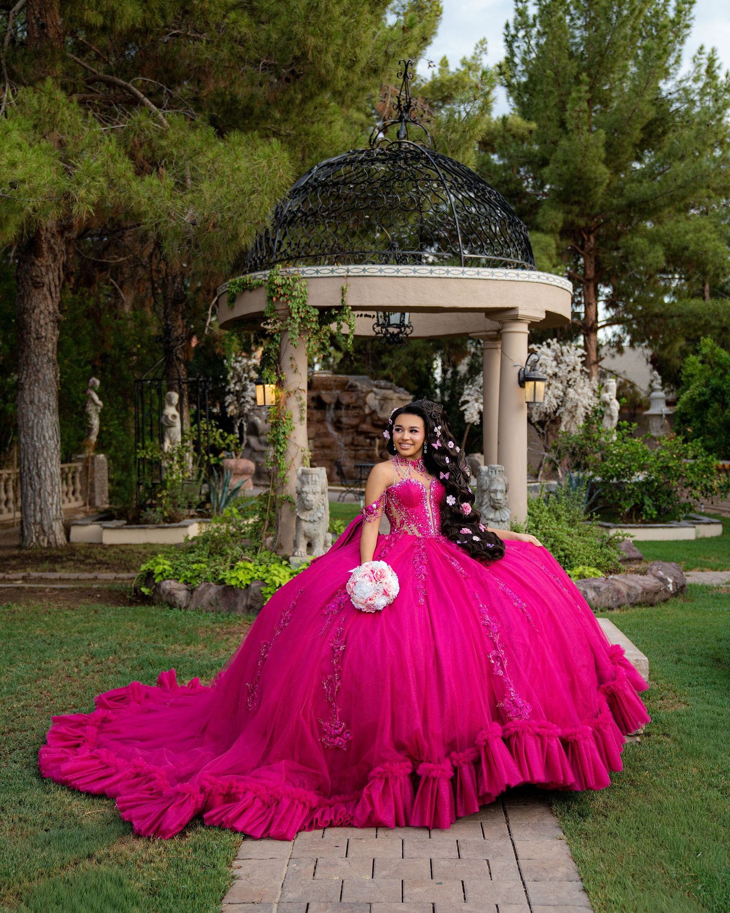 Young woman in a vibrant hot pink quinceañera ball gown holding a bouquet in a lush garden gazebo.