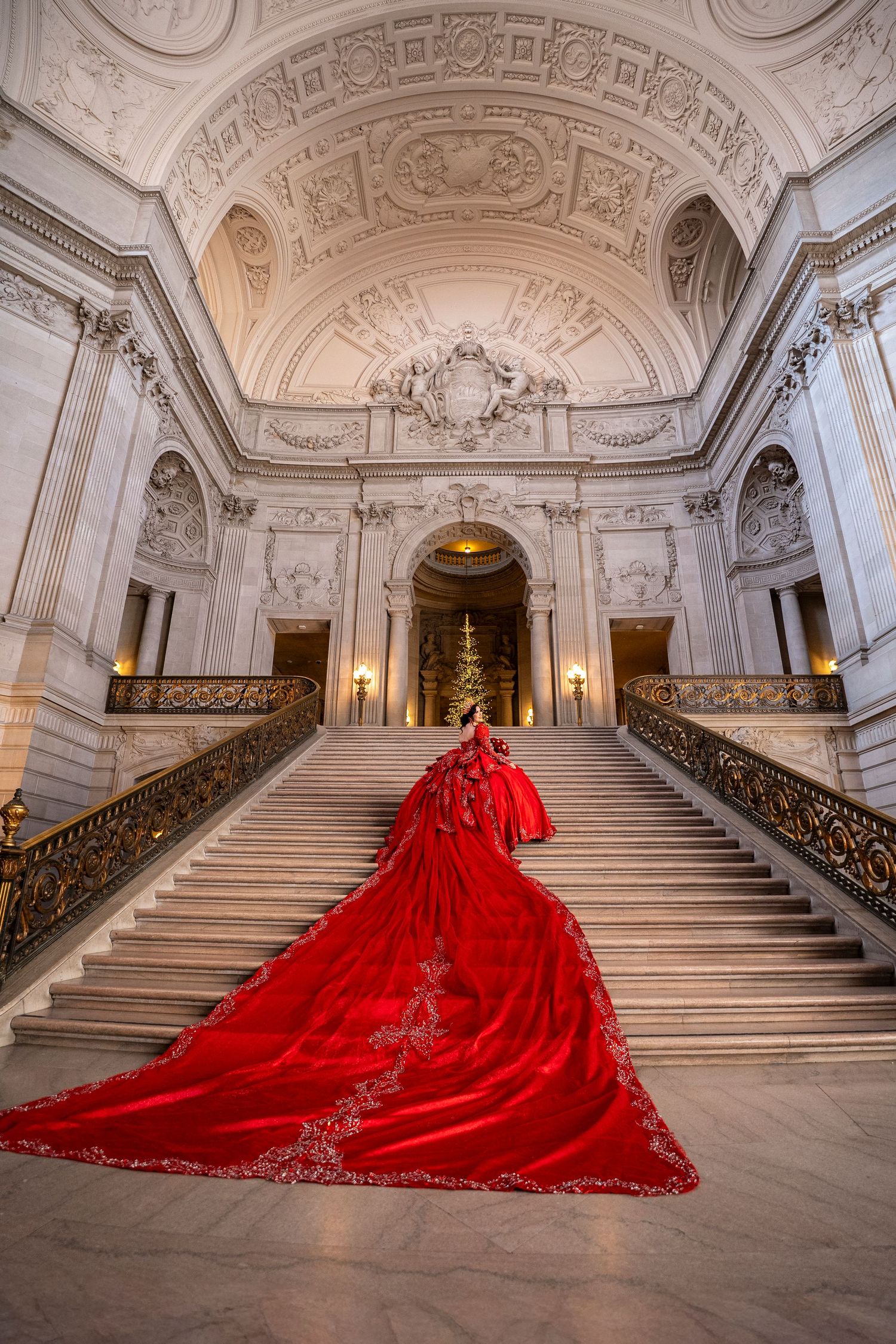Rear view of woman in red embroidered ball gown with dramatic train ascending ornate marble stairs.