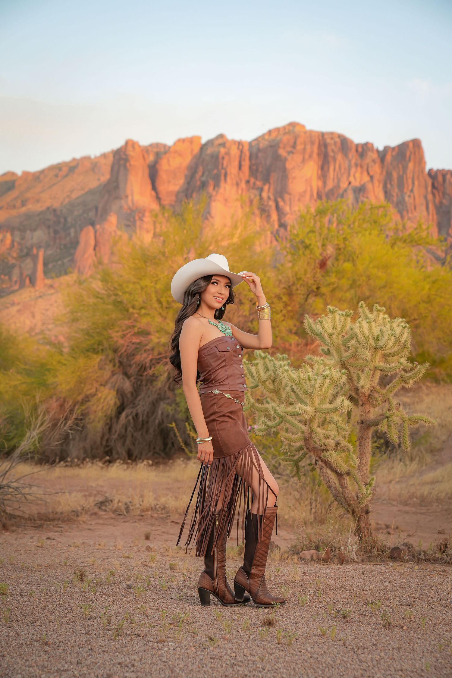 Woman in brown fringe dress and white cowboy hat poses before red rock mountains and cacti in Arizona desert at sunset.