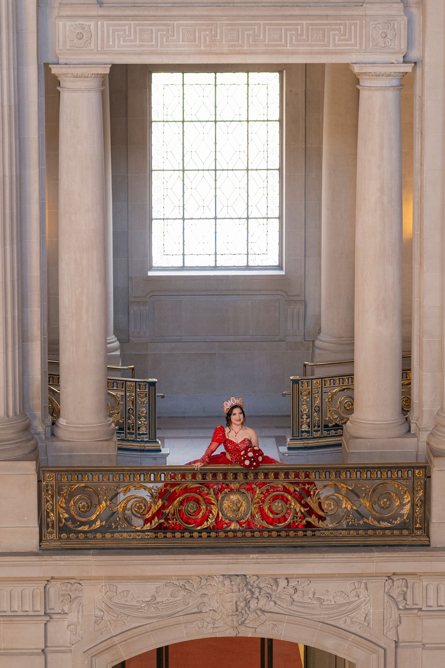 Woman in red gown and crown poses on ornate golden balcony inside grand neoclassical building.