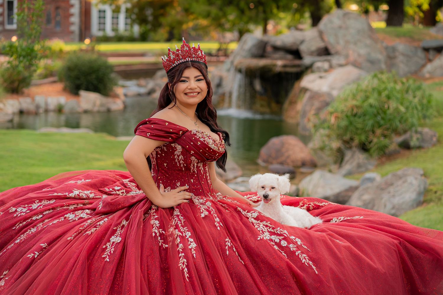 Young woman in red quinceañera ball gown with floral embroidery and crown poses with white dog by garden waterfall.