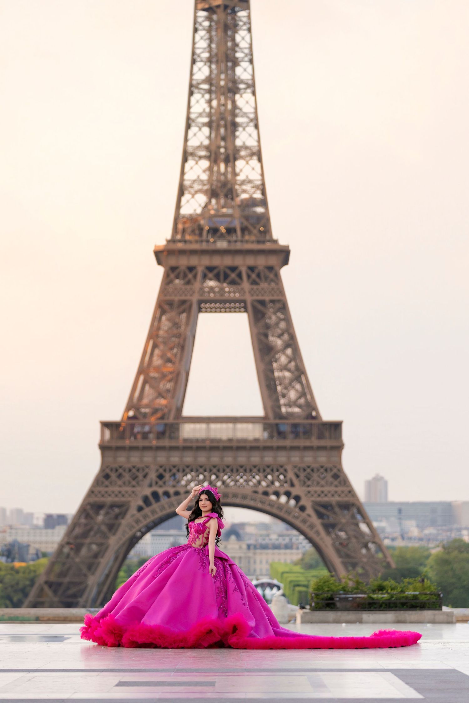 Quinceañera in a vibrant pink ball gown with long train poses in front of the Eiffel Tower in Paris.