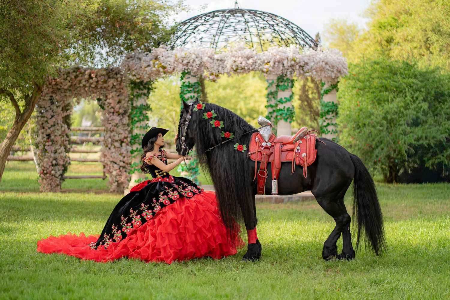 Girl in red and black quinceañera gown poses with black horse near floral arch in lush green garden.