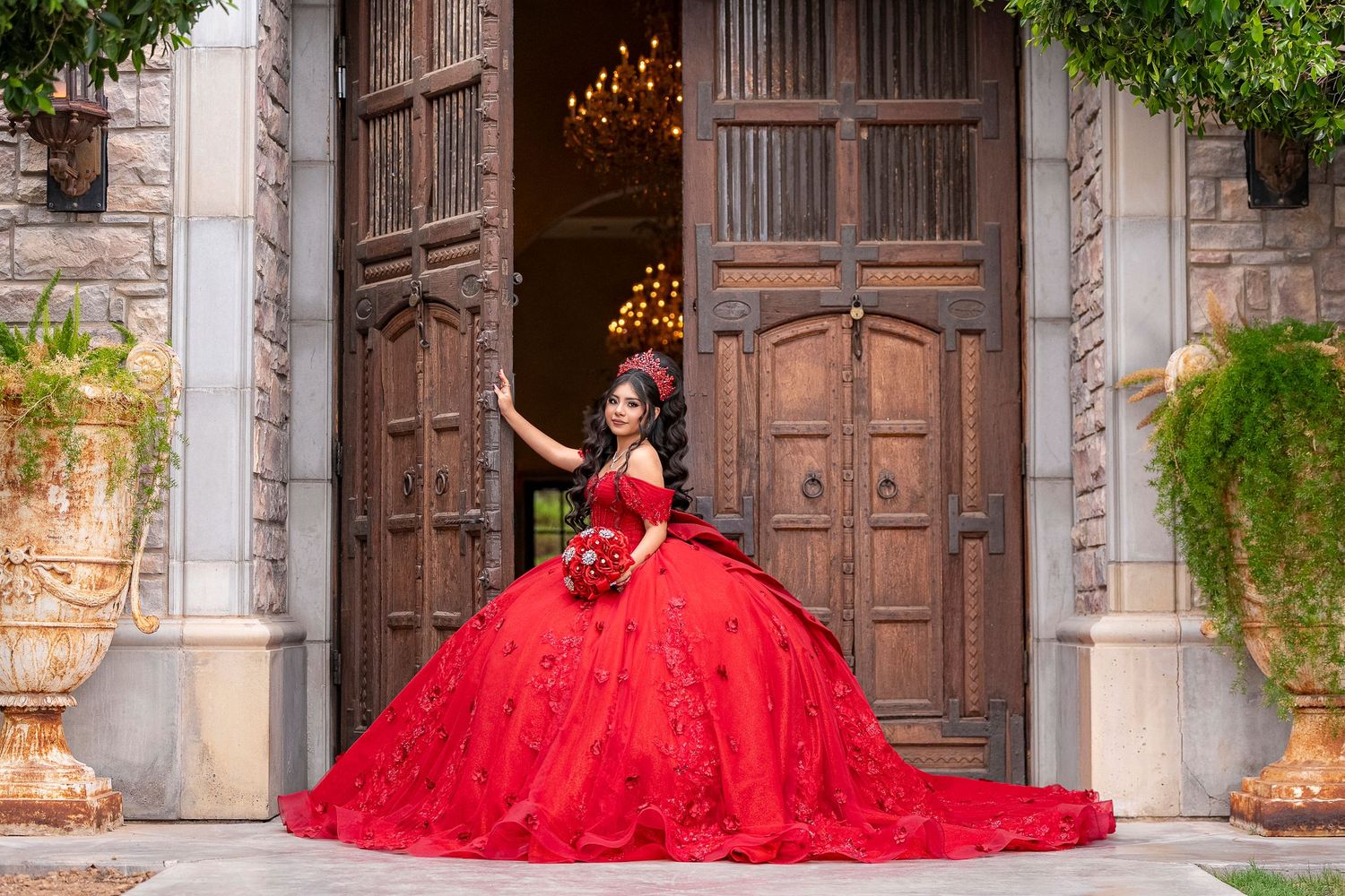 Young woman in a stunning red ball gown quinceañera dress poses at ornate wooden doors of a stone mansion.