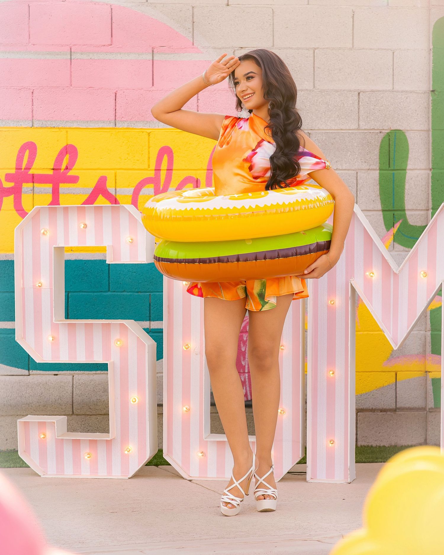 Woman in colorful romper holding inflatable hamburger float beside large marquee SUN letters at a vibrant summer party.