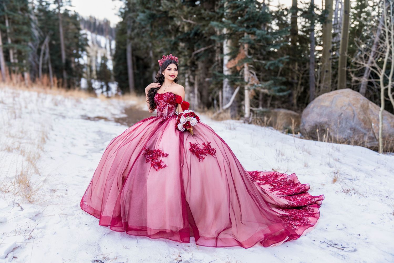 Woman in pink ballgown with red floral appliques poses in snowy winter landscape surrounded by pine trees.