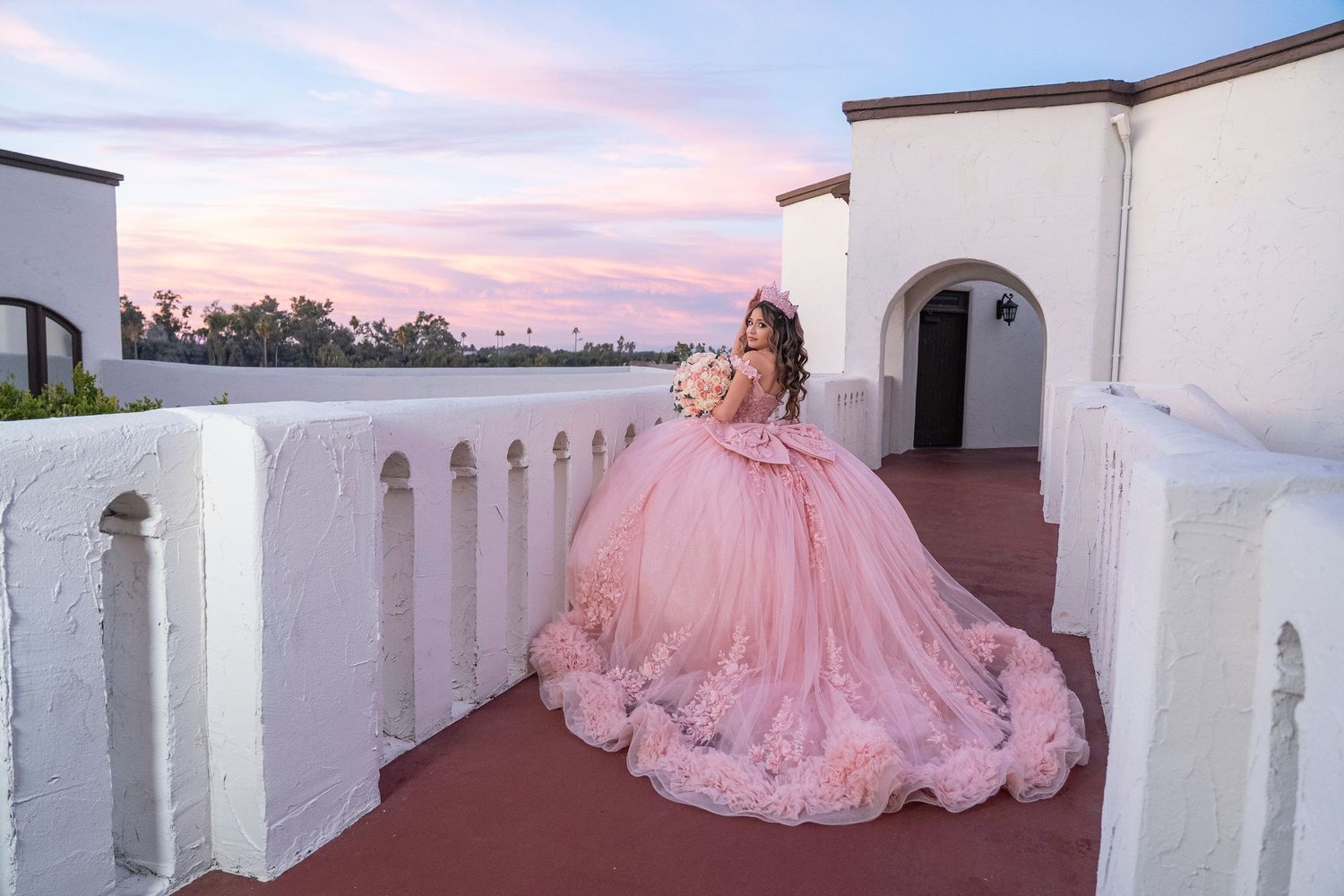 Young woman in a pink ball gown quinceañera dress poses on a rooftop balcony at sunset.