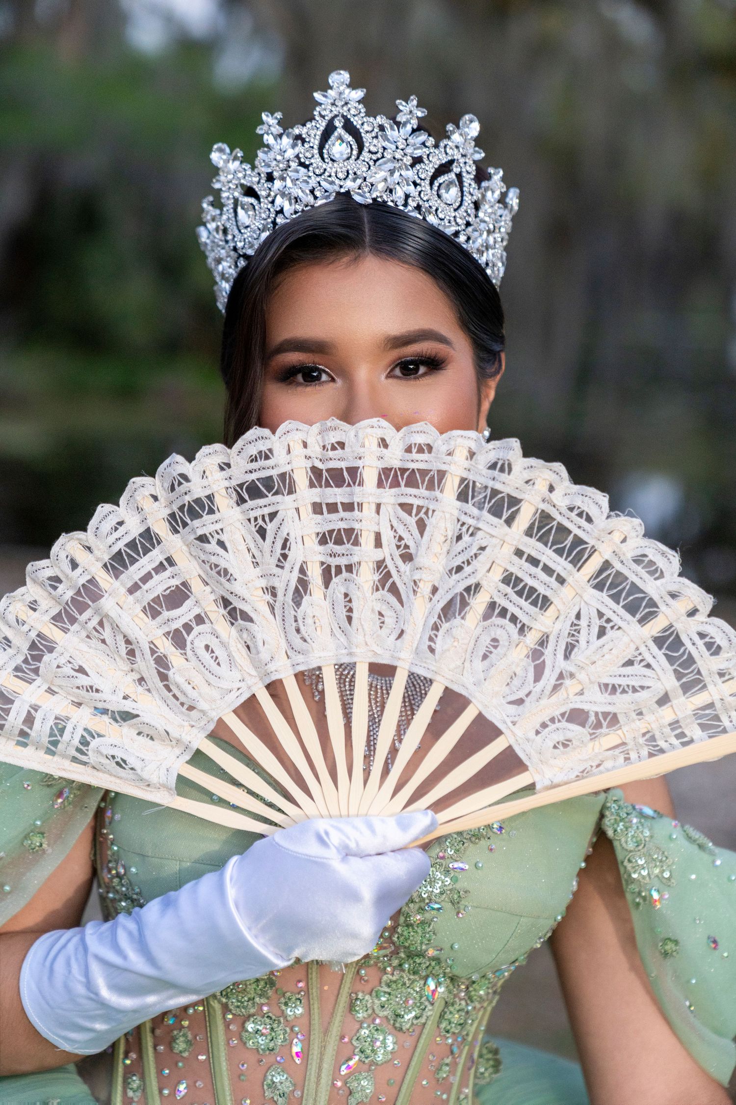 Young woman in green gown, diamond tiara, white gloves, holding elegant lace fan at quinceañera celebration.