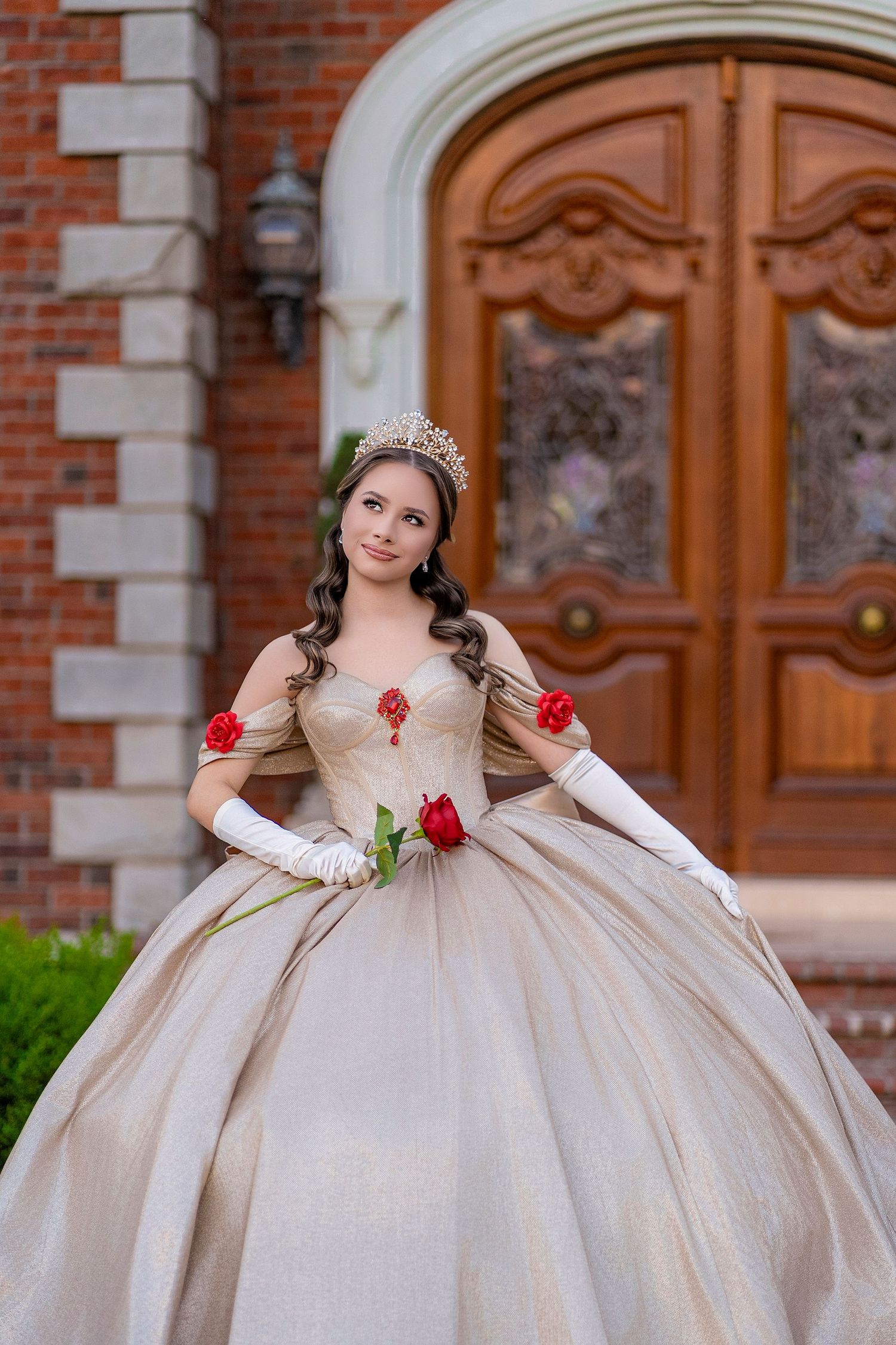 Young woman in gold ball gown with crown and red roses poses before ornate wooden doors for quinceañera.