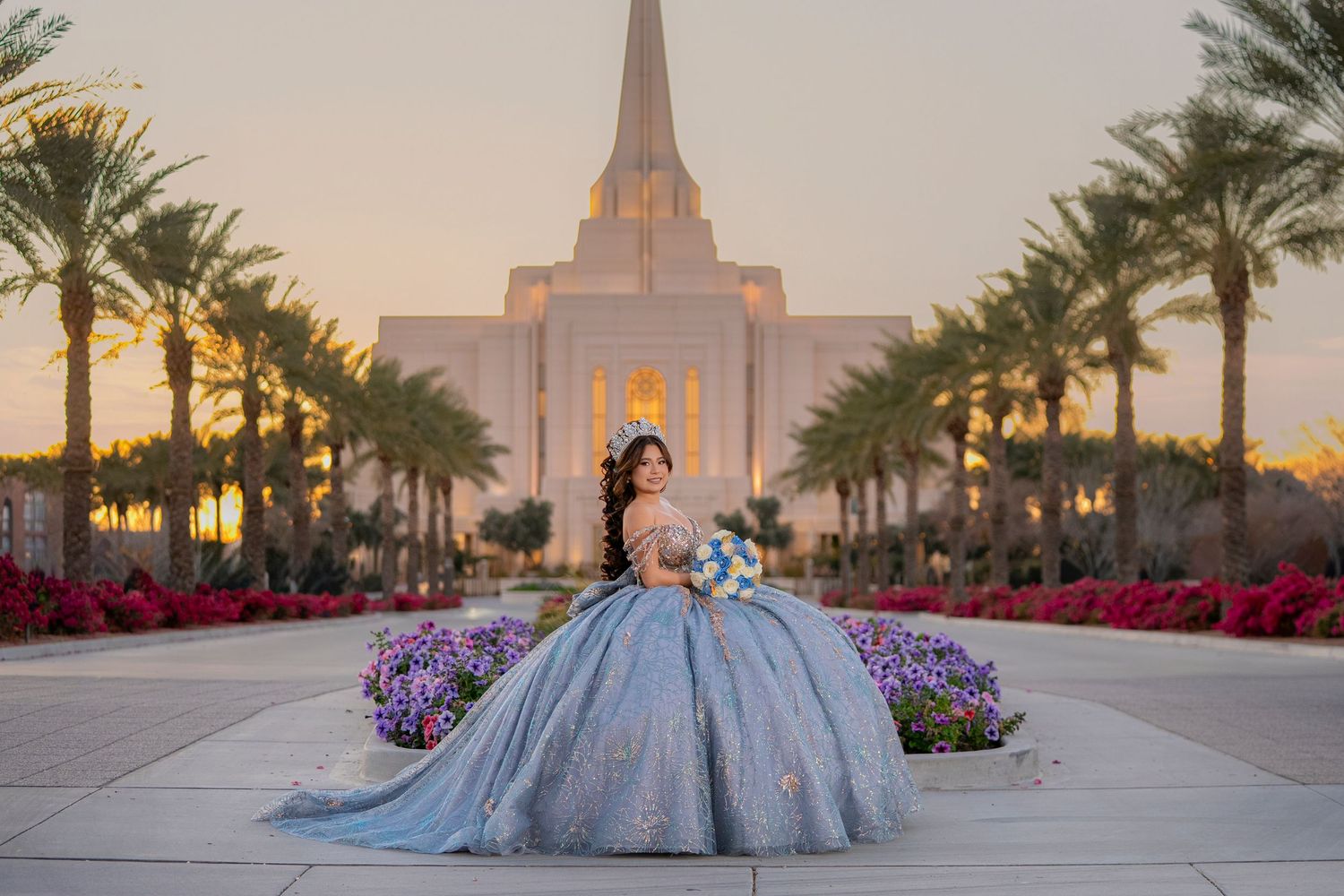 Young woman in a blue ball gown poses at a white temple surrounded by palm trees and colorful flowers at sunset.