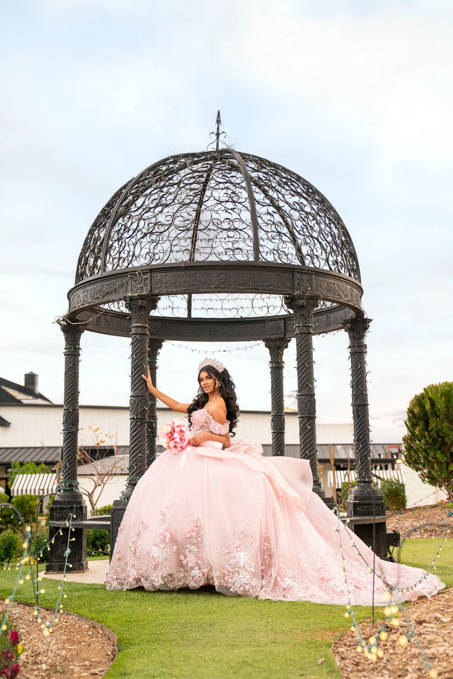 Young woman in a pink ball gown poses under an ornate iron gazebo in a garden for her quinceañera.
