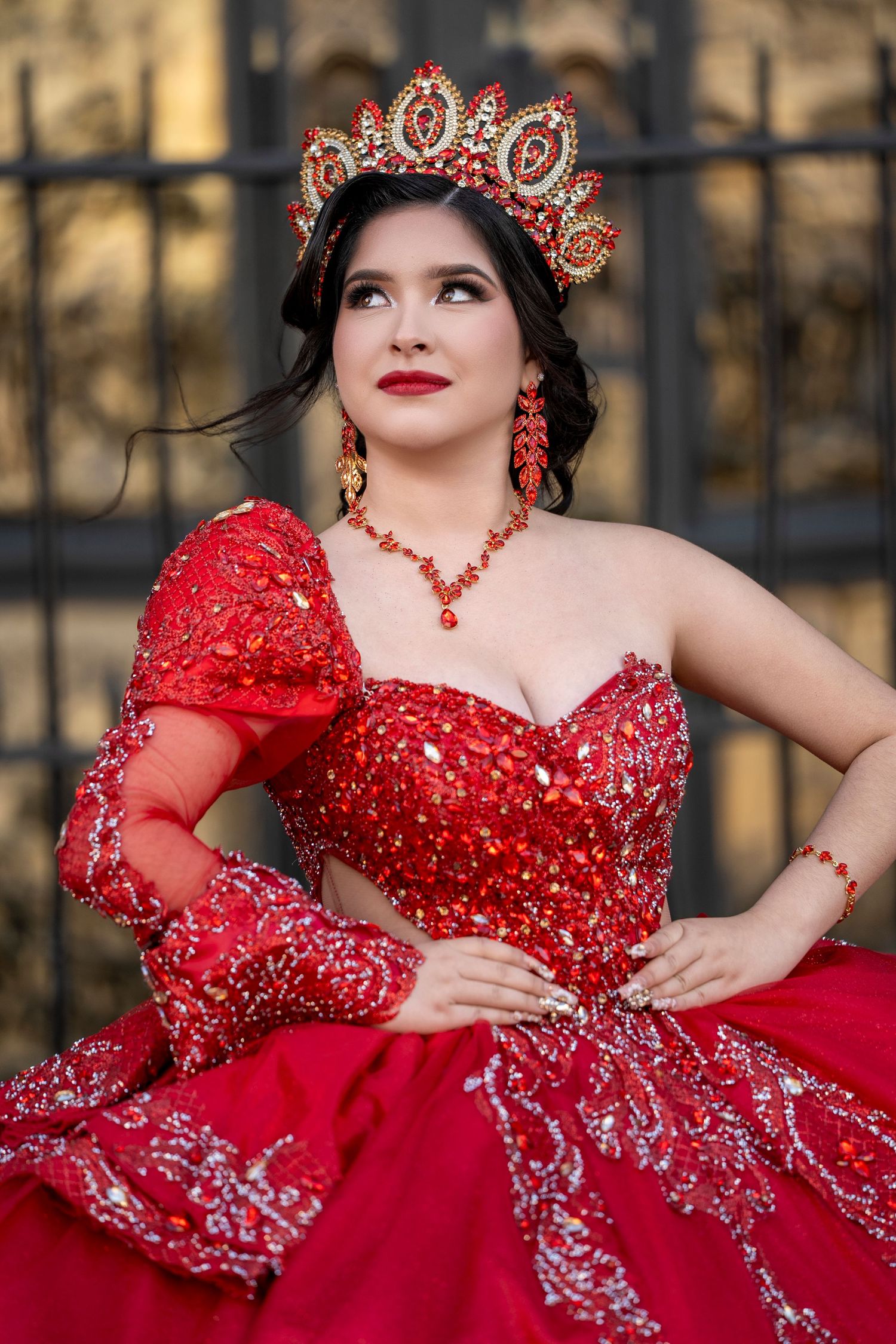 Young woman in a red sequined quinceañera gown with matching crown, jewelry, and gloves poses elegantly outdoors.