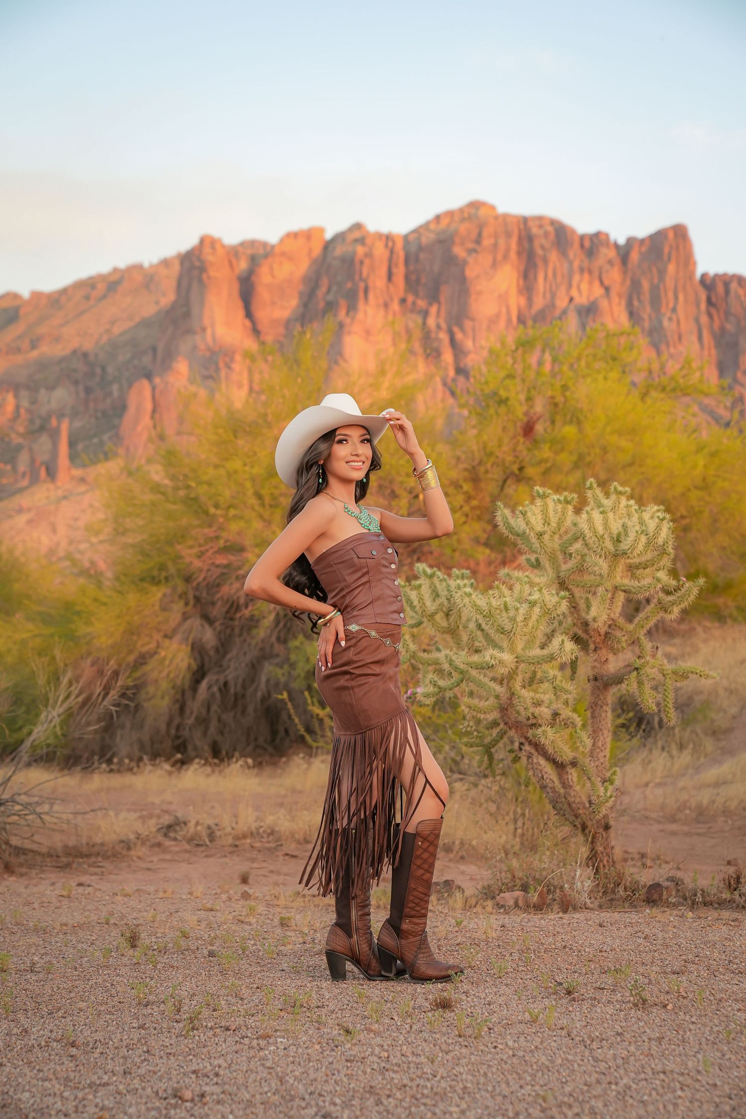 Woman in brown fringe dress, white cowboy hat, and boots poses in Arizona desert with red rock mountains and cactus.