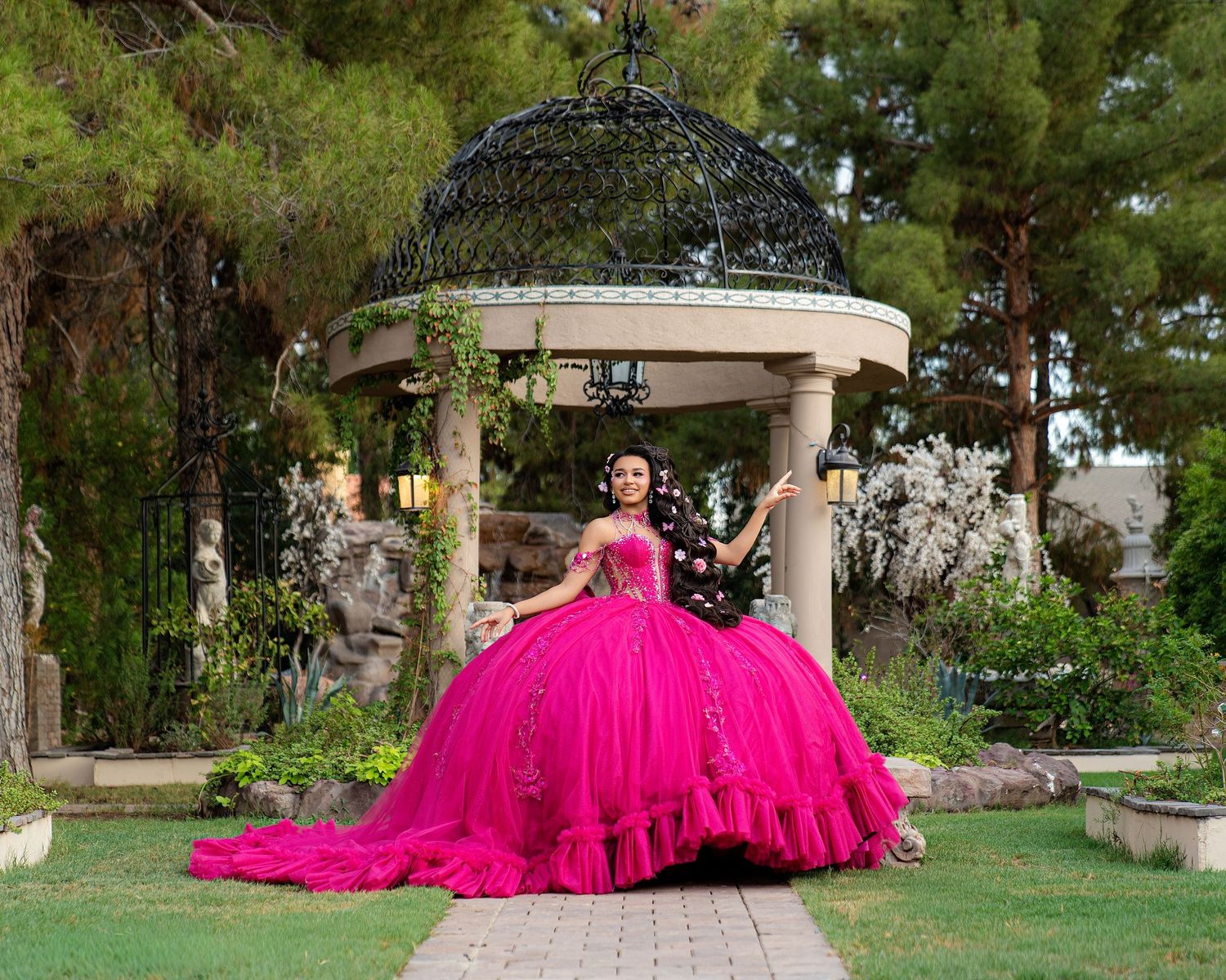 Young woman in a vibrant hot pink ball gown poses in a lush garden gazebo setting for a quinceañera celebration.