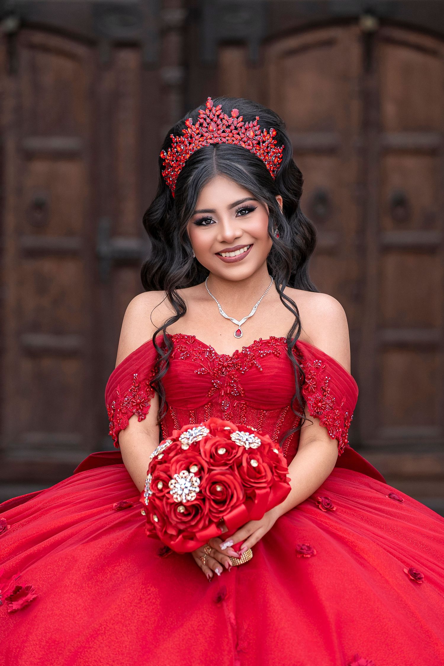 Young Latina in red quinceañera ball gown with crown holds red rose bouquet, smiling before wooden doors.