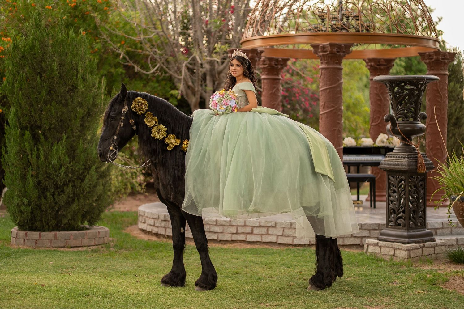 Young woman in green quinceañera gown poses on black horse decorated with yellow flowers near brick gazebo.