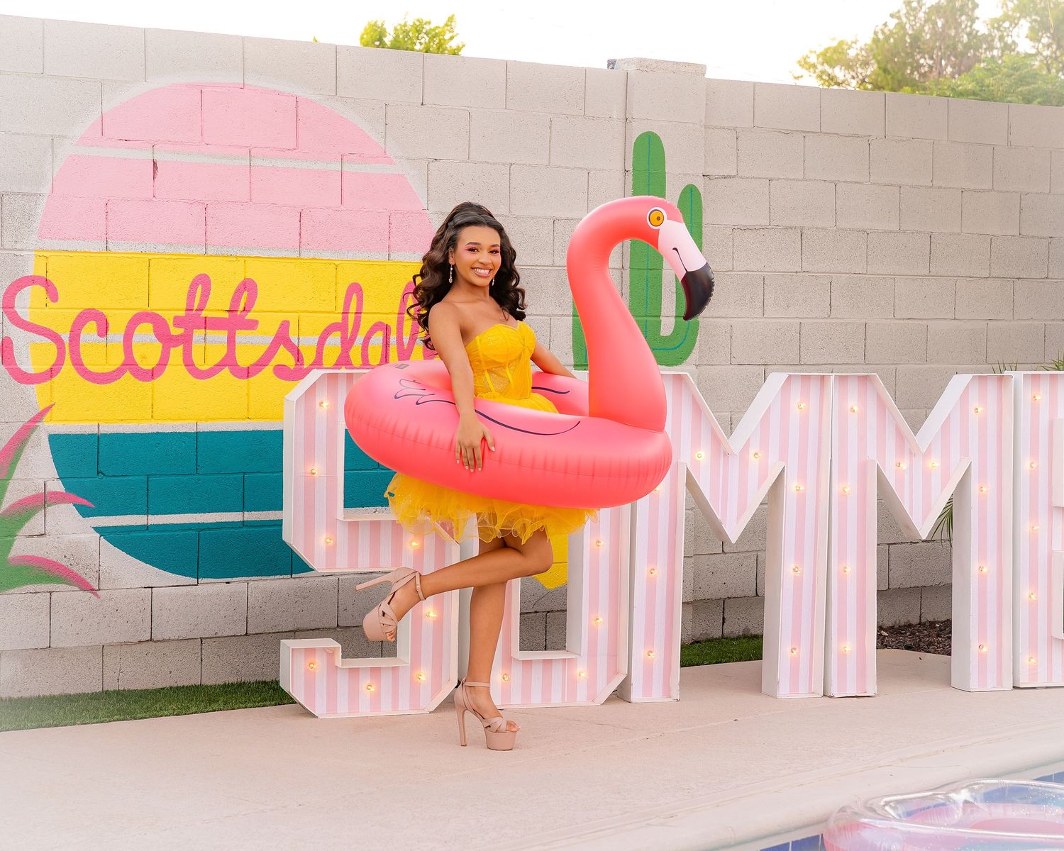 Woman in yellow dress holding pink flamingo pool float poses by Scottsdale Summer mural with giant lit letters.