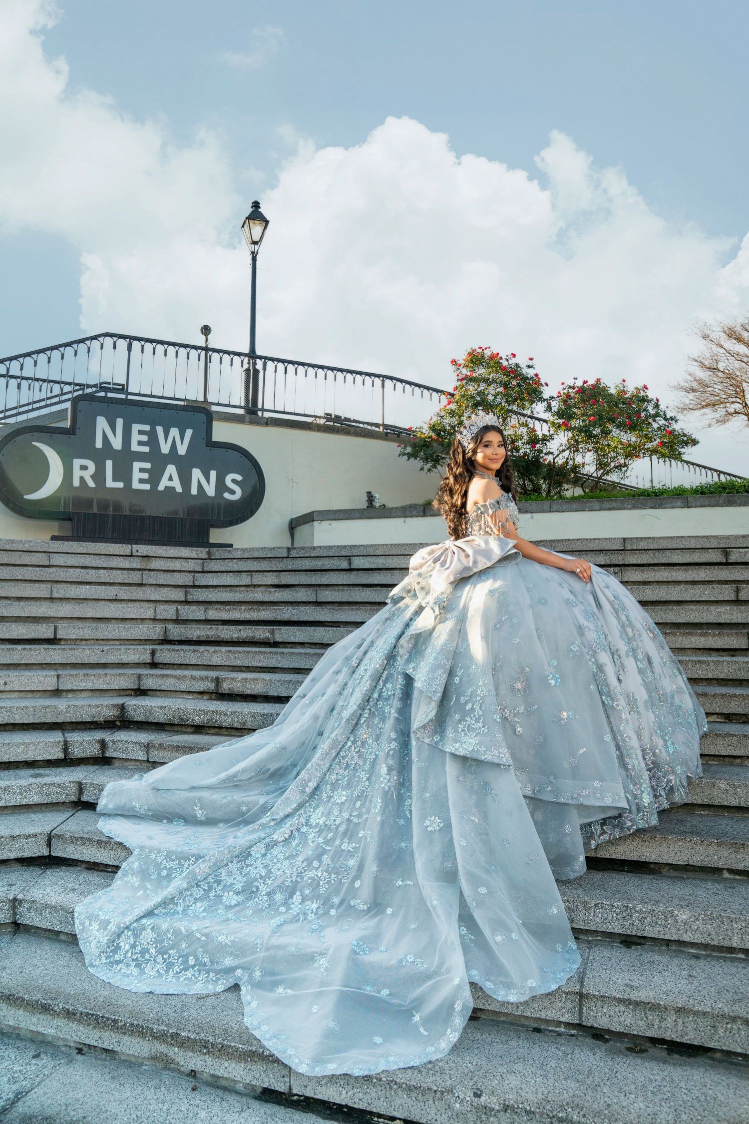 A woman in a stunning light blue ball gown poses on stone steps beneath a New Orleans sign under a cloudy sky.
