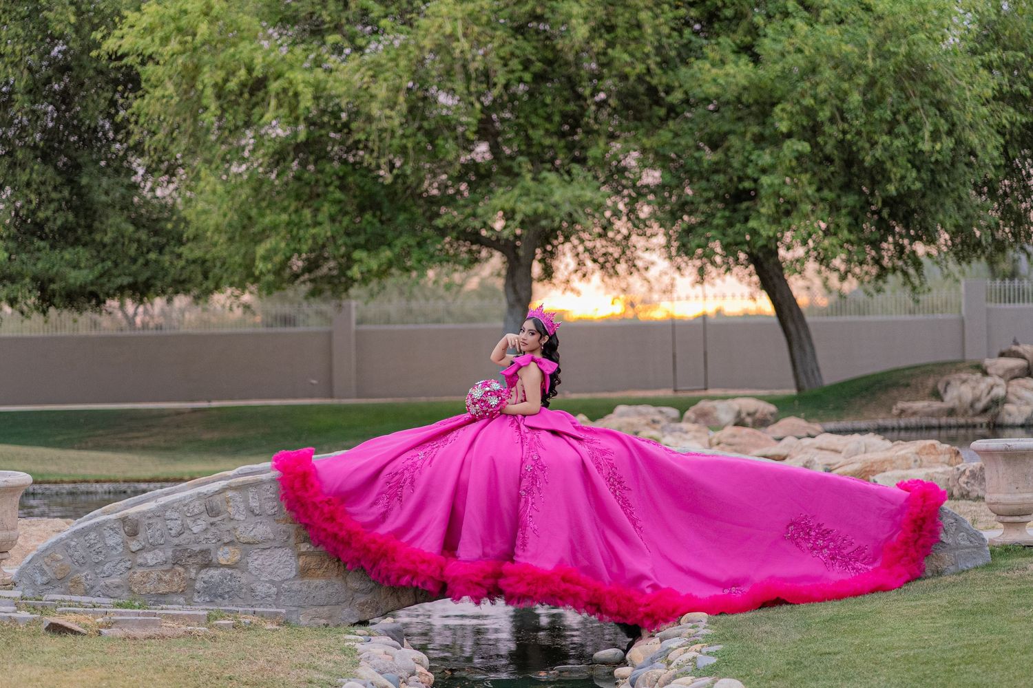 Elegant quinceañera in a vibrant hot pink ball gown twirls near a pond at sunset surrounded by trees.