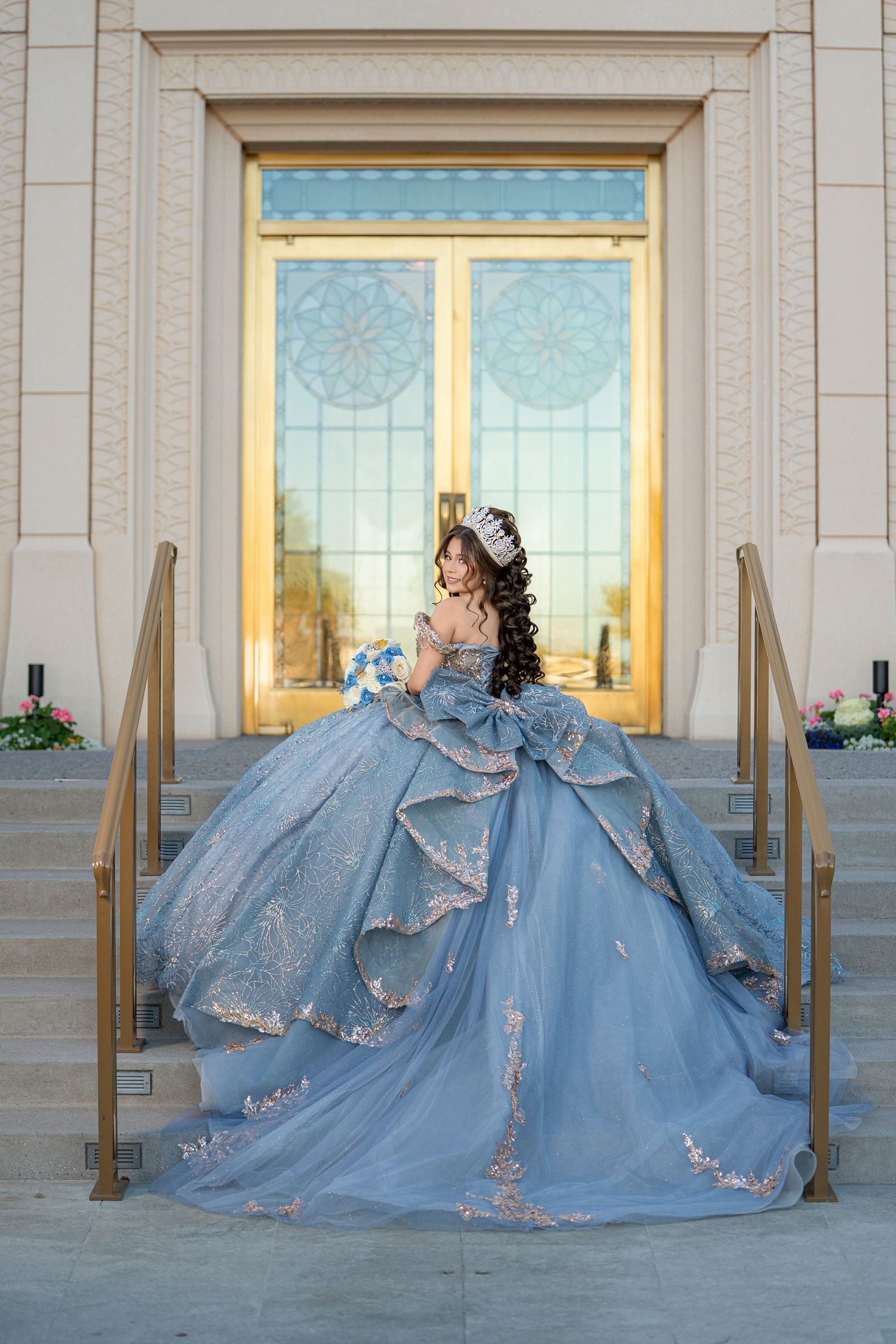 Young woman in a stunning blue ball gown with floral lace details poses on elegant building steps.