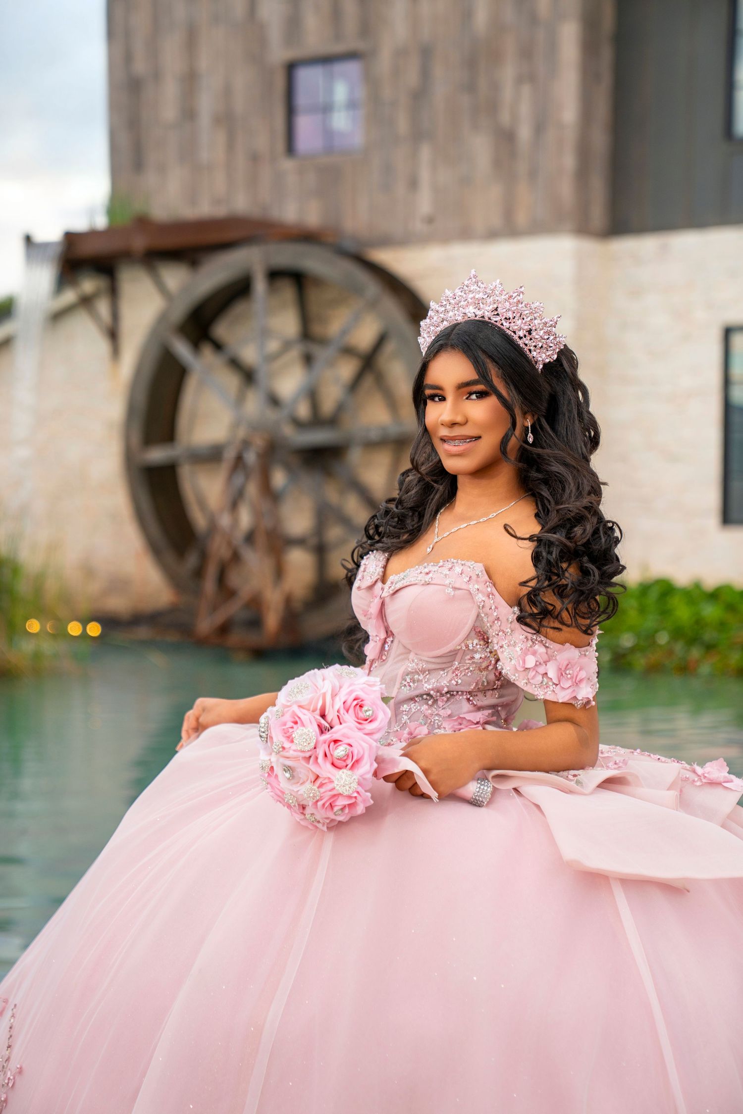Young woman in pink quinceañera ball gown with floral crown holds rose bouquet near rustic watermill backdrop.