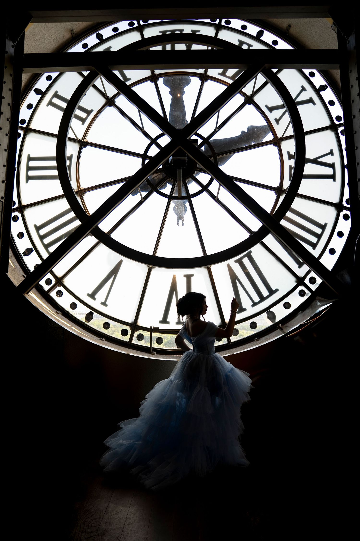 Silhouette of a quinceañera in a blue ball gown posed before a massive ornate clock face in Paris.