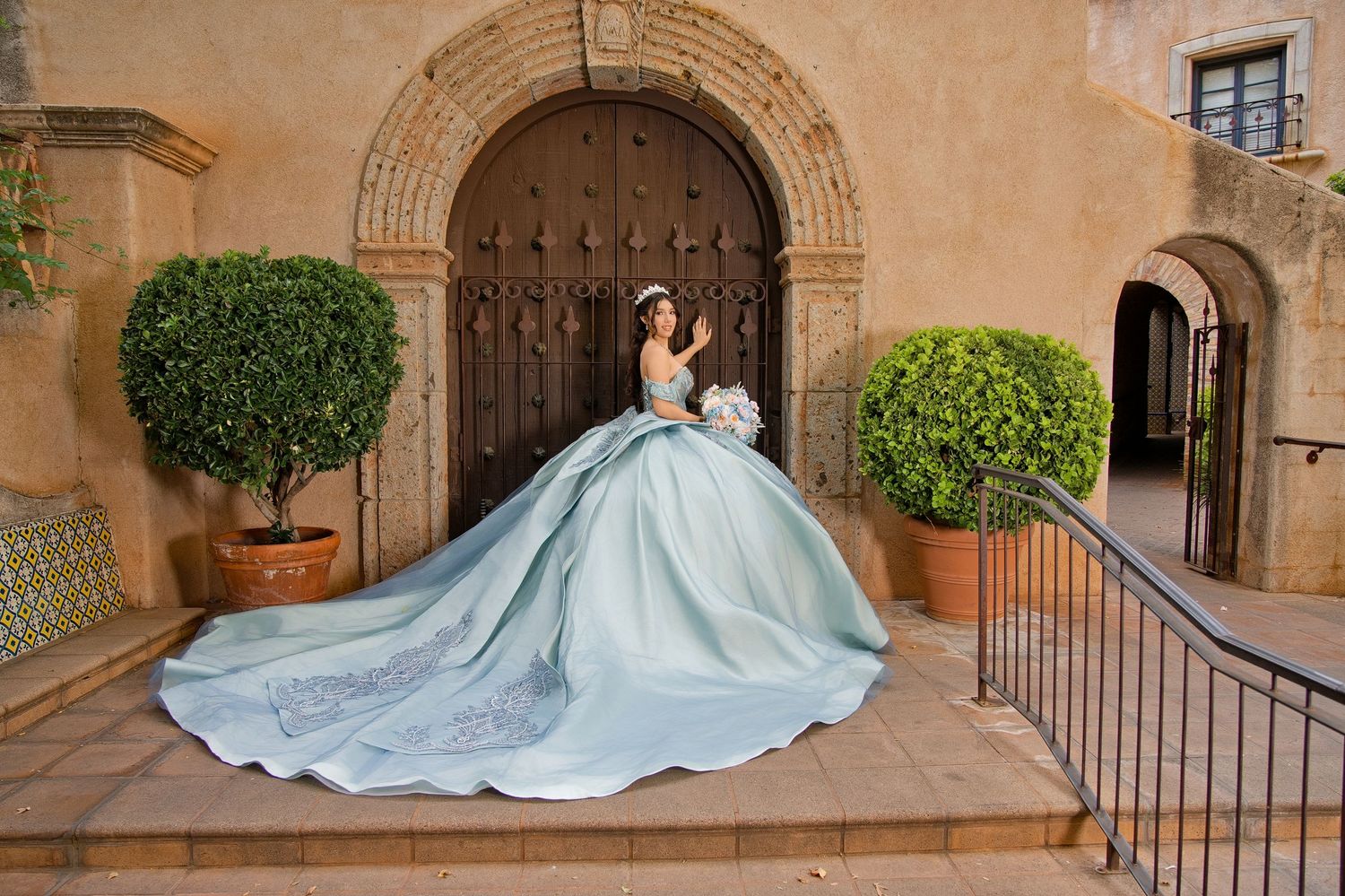 Young woman in elegant light blue quinceañera ball gown with long train poses on stone steps by ornate wooden doors.