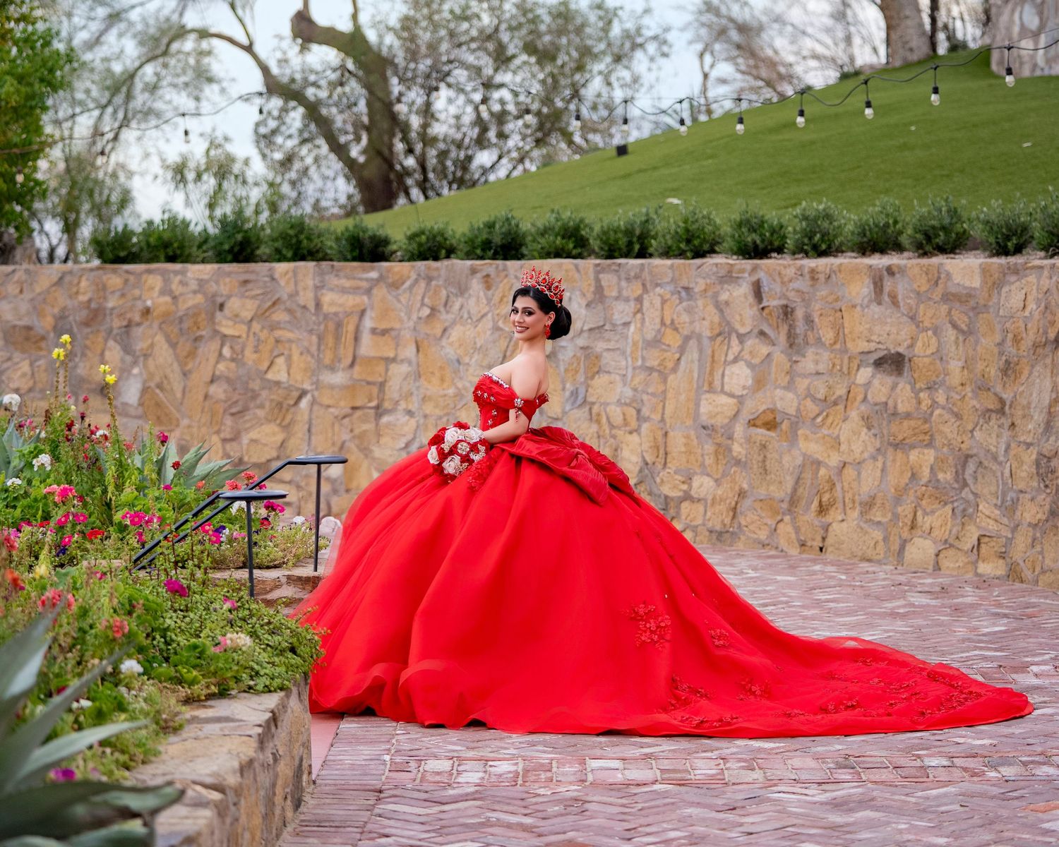 Young woman in a stunning red ball gown with long train poses in a garden for a quinceañera photo.