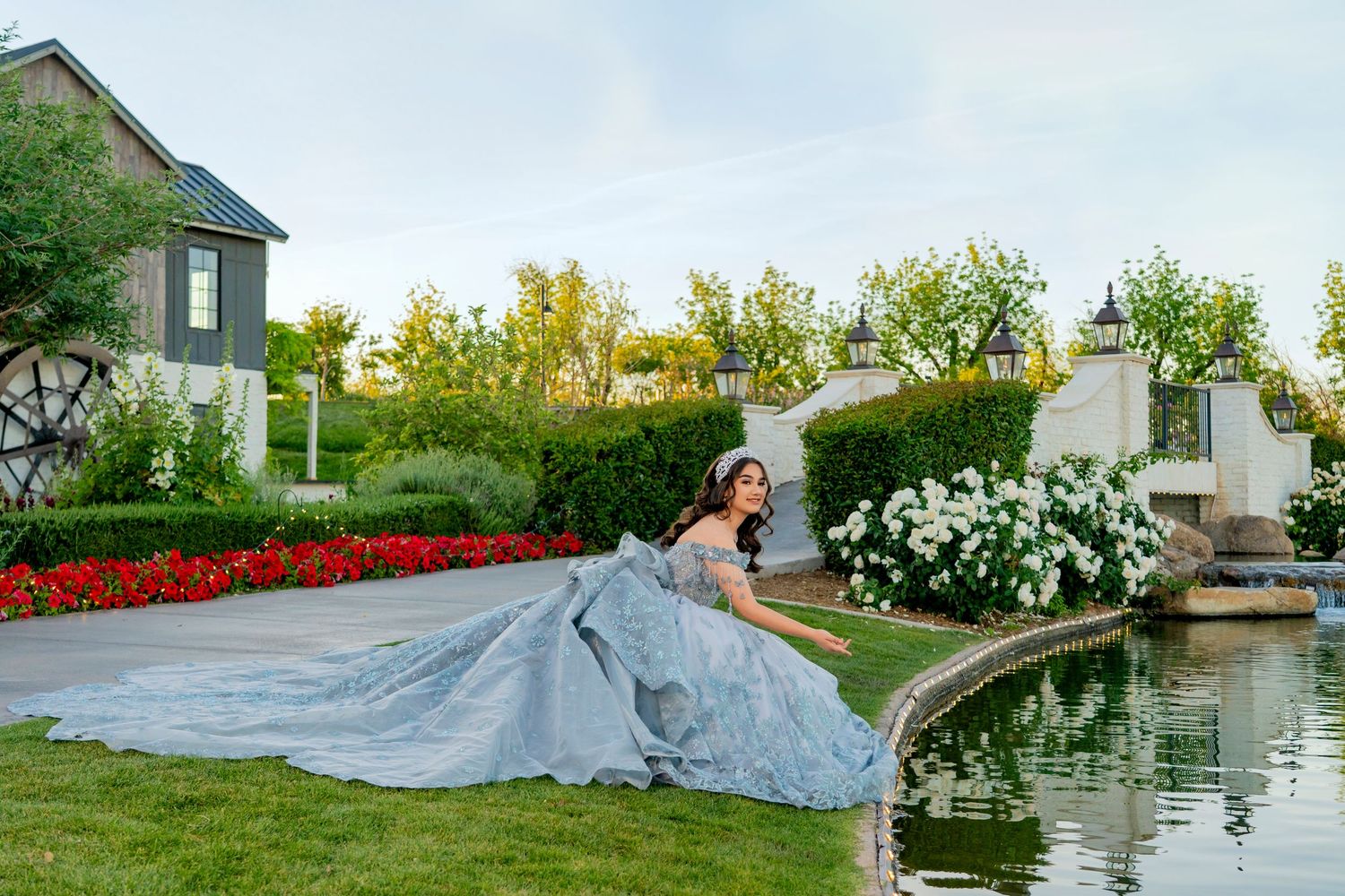 A woman in a stunning silver ball gown poses gracefully by a garden pond surrounded by lush greenery and colorful flowers.