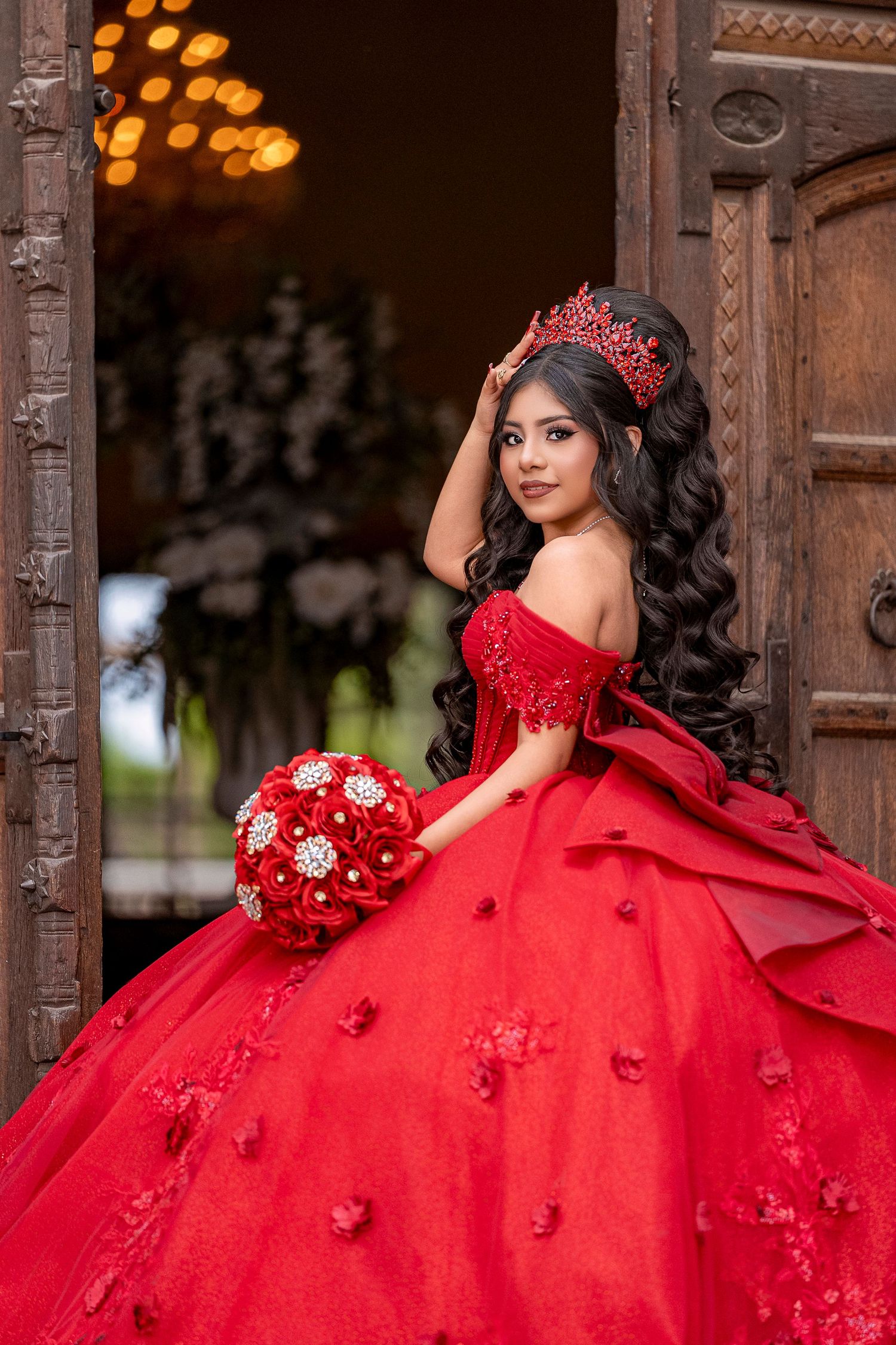 Young woman in red ball gown with crown and bouquet posing in ornate doorway for quinceañera.