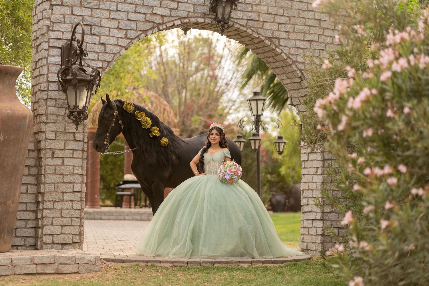 Young woman in mint green quinceañera ball gown poses under stone arch with black horse in garden setting.
