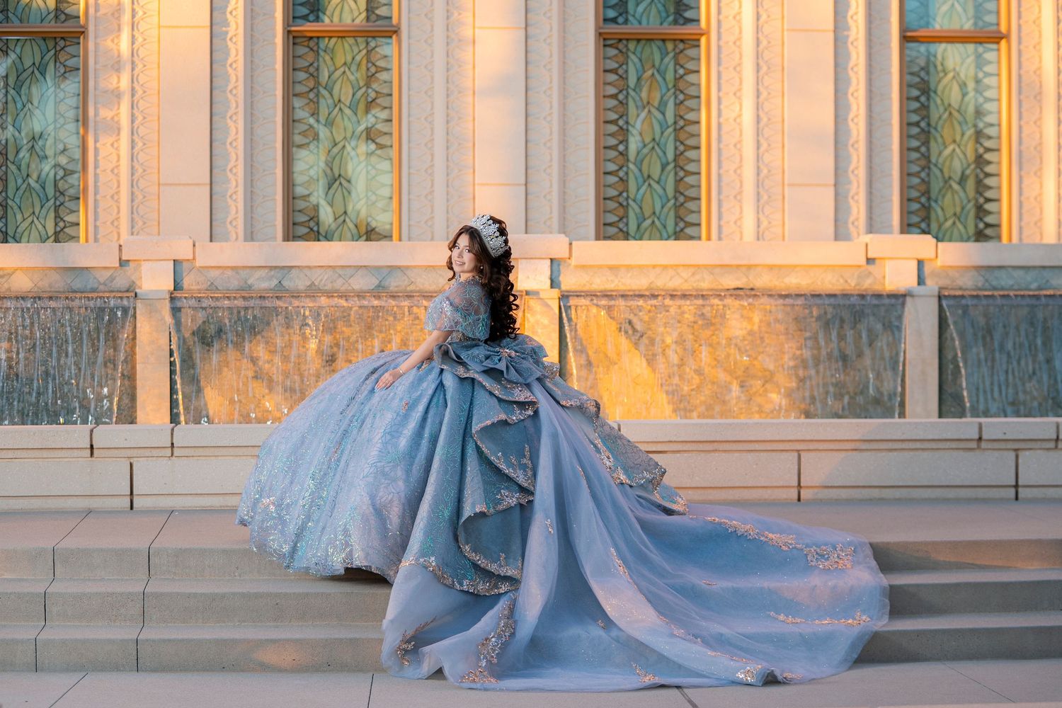 Woman in elegant blue ball gown with long train poses on grand steps of ornate building at golden hour.