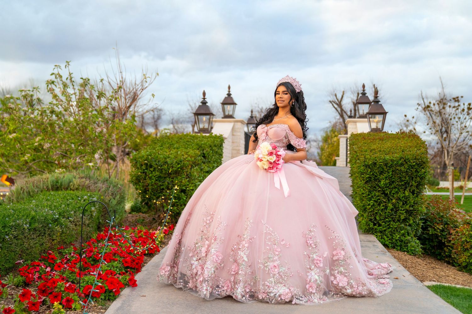 Young woman in pink quinceañera ball gown with floral appliqués poses in garden with red flowers and decorative lanterns.