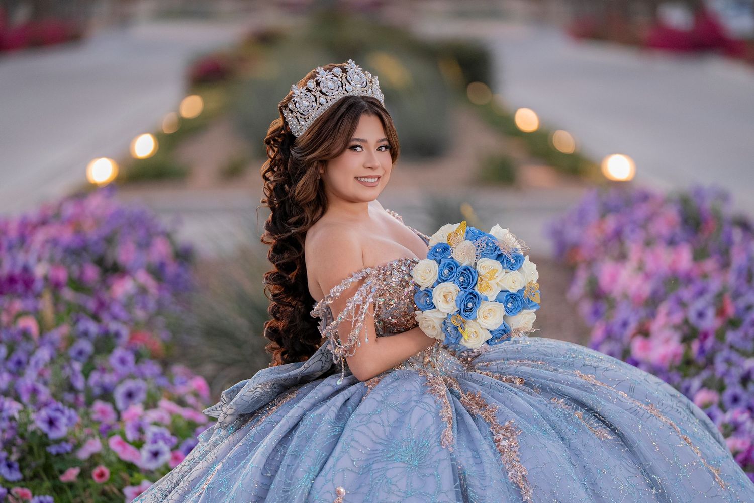 Young woman in blue quinceañera ball gown with crown and floral bouquet poses in blooming garden.
