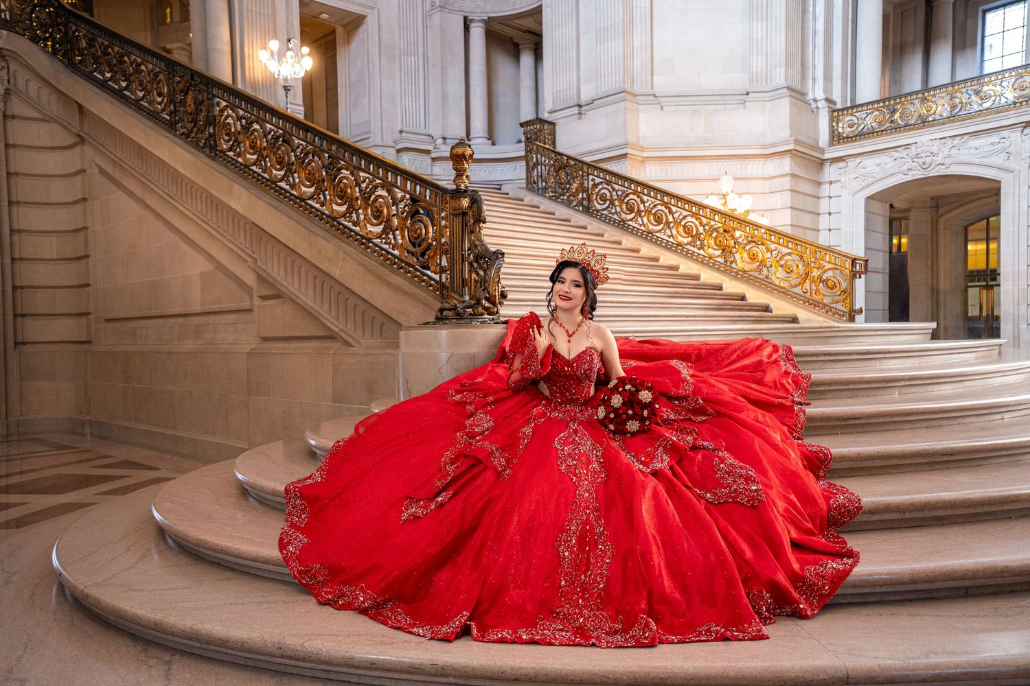 Young woman in a stunning red ball gown with gold embroidery poses on grand marble staircase for quinceañera photos.