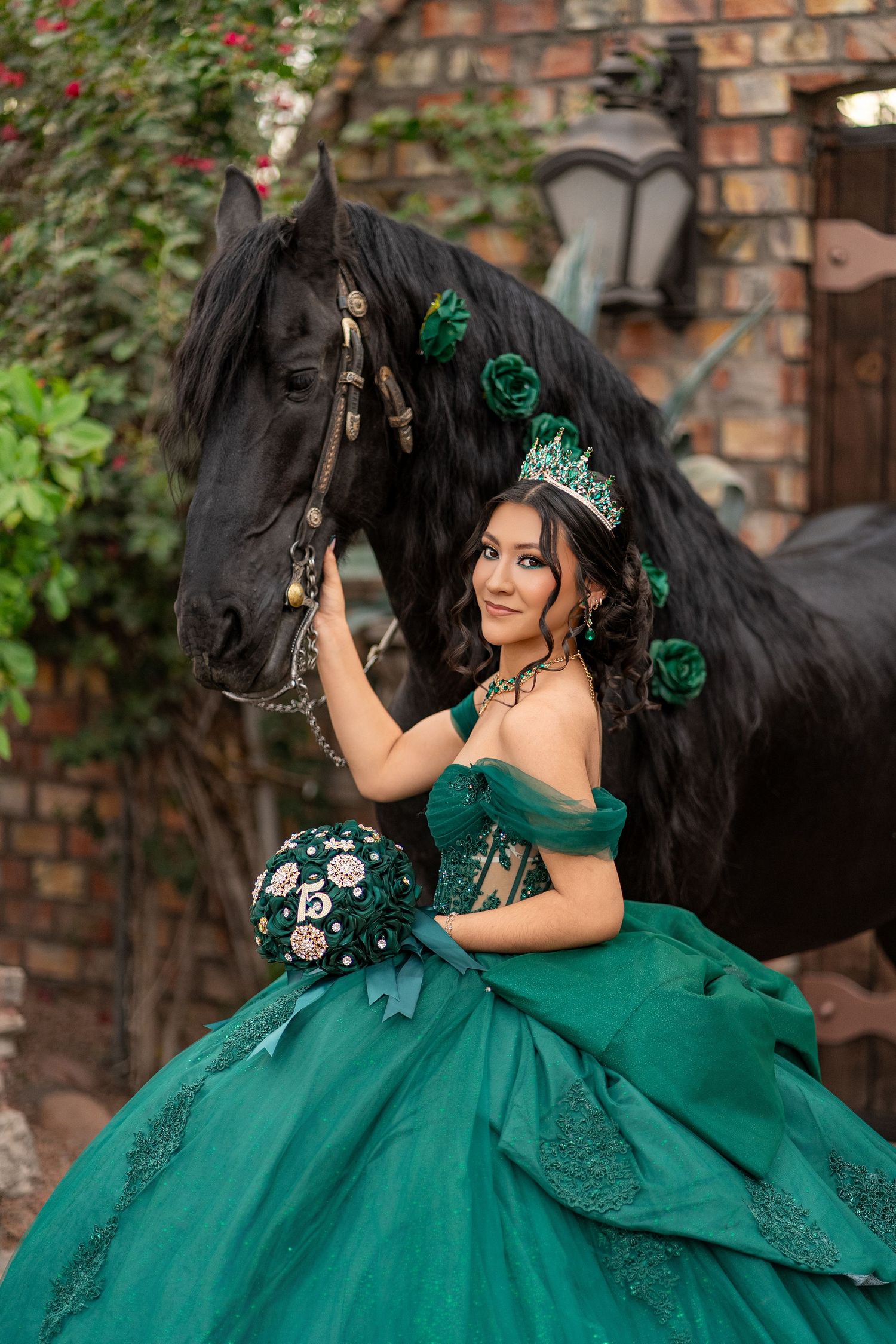 Young woman in emerald green quinceañera gown and crown poses with black horse in a fairytale garden setting.