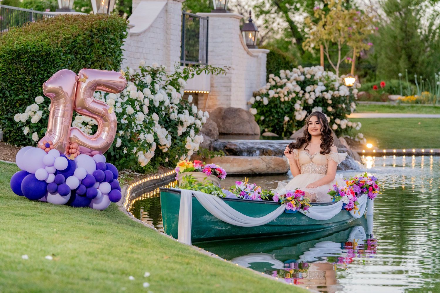 A girl celebrates her quinceañera sitting in a decorated boat on a pond, with number 15 balloons nearby.