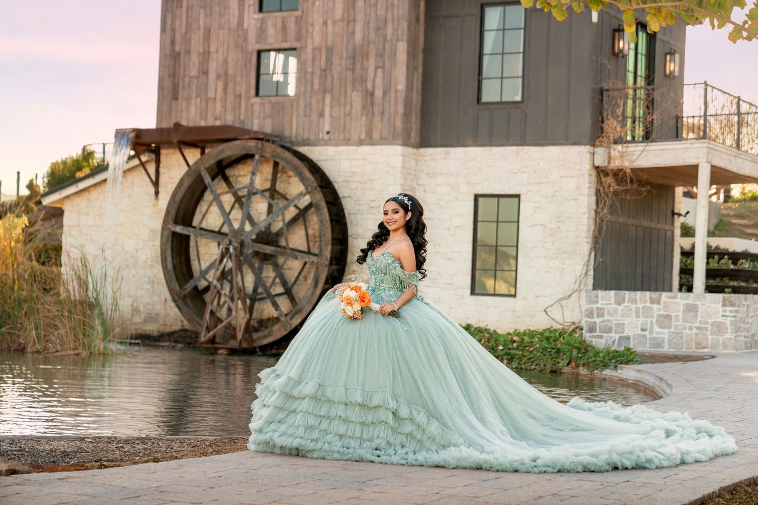 Young woman in a mint green quinceañera ball gown poses by a water wheel holding an orange bouquet.
