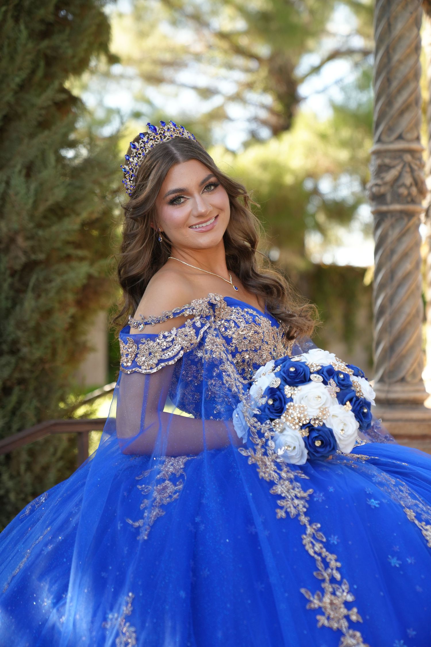 Smiling young woman in royal blue quinceañera gown with gold embroidery and tiara holds floral bouquet.