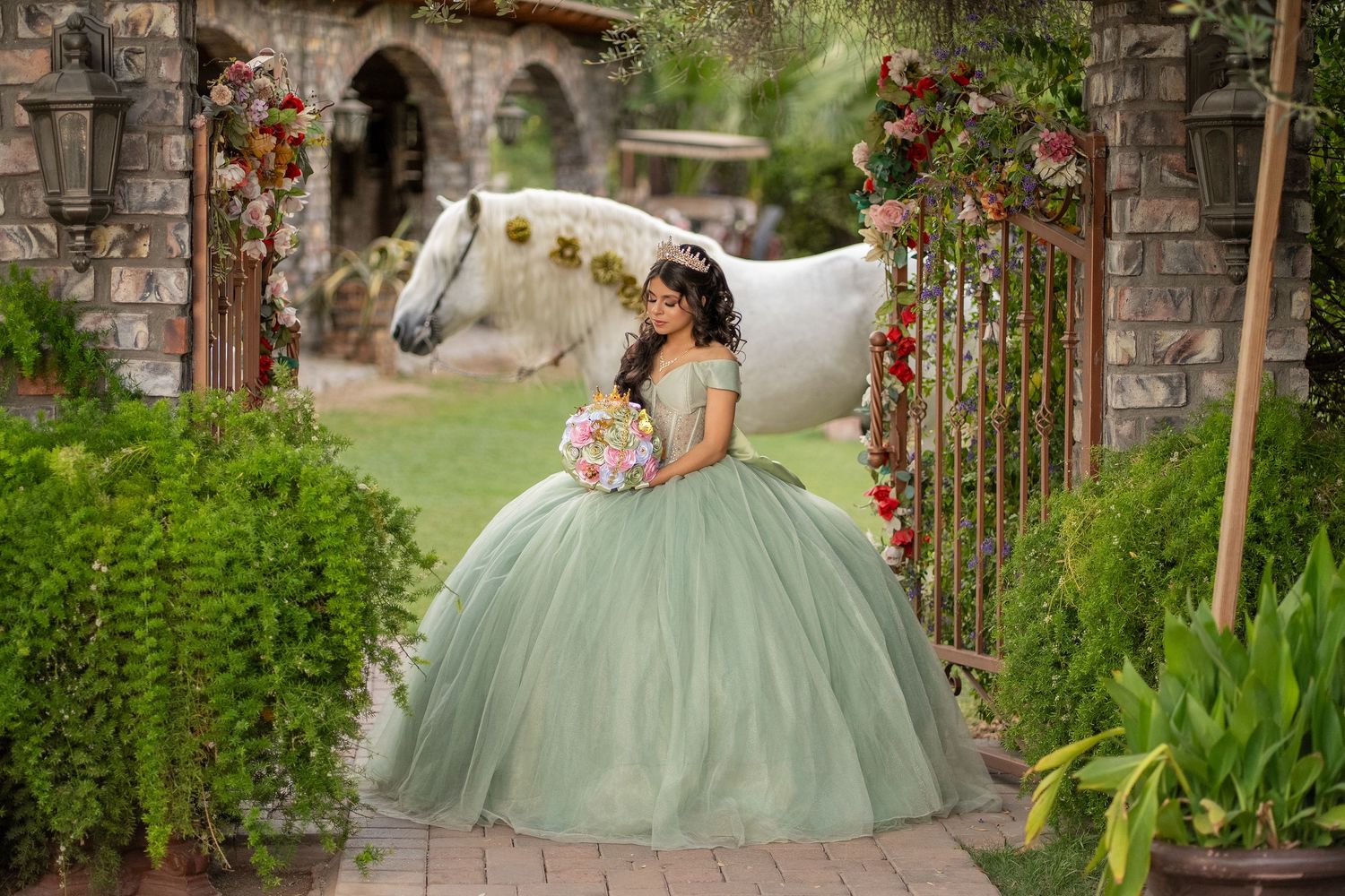 Young woman in sage green quinceañera ball gown poses with white horse amid floral arch in garden setting.