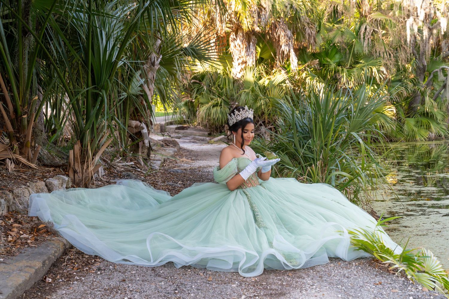 Young woman in mint green quinceañera ball gown with tiara poses gracefully in a lush tropical garden setting.