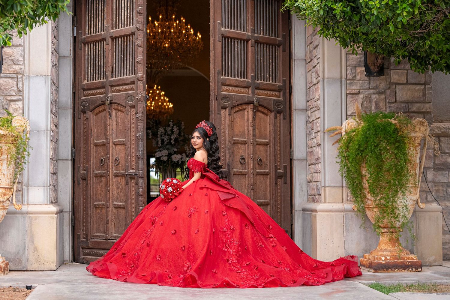 Young woman in a stunning red ball gown quinceañera dress poses at ornate wooden doors of an elegant venue.