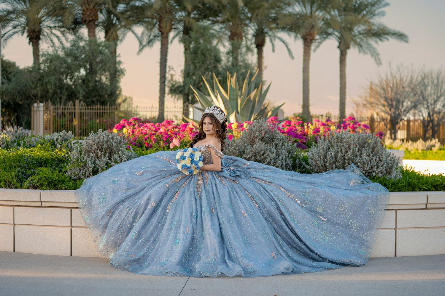 Young woman in a sparkling blue ball gown and crown poses gracefully outdoors with colorful flowers and palm trees.