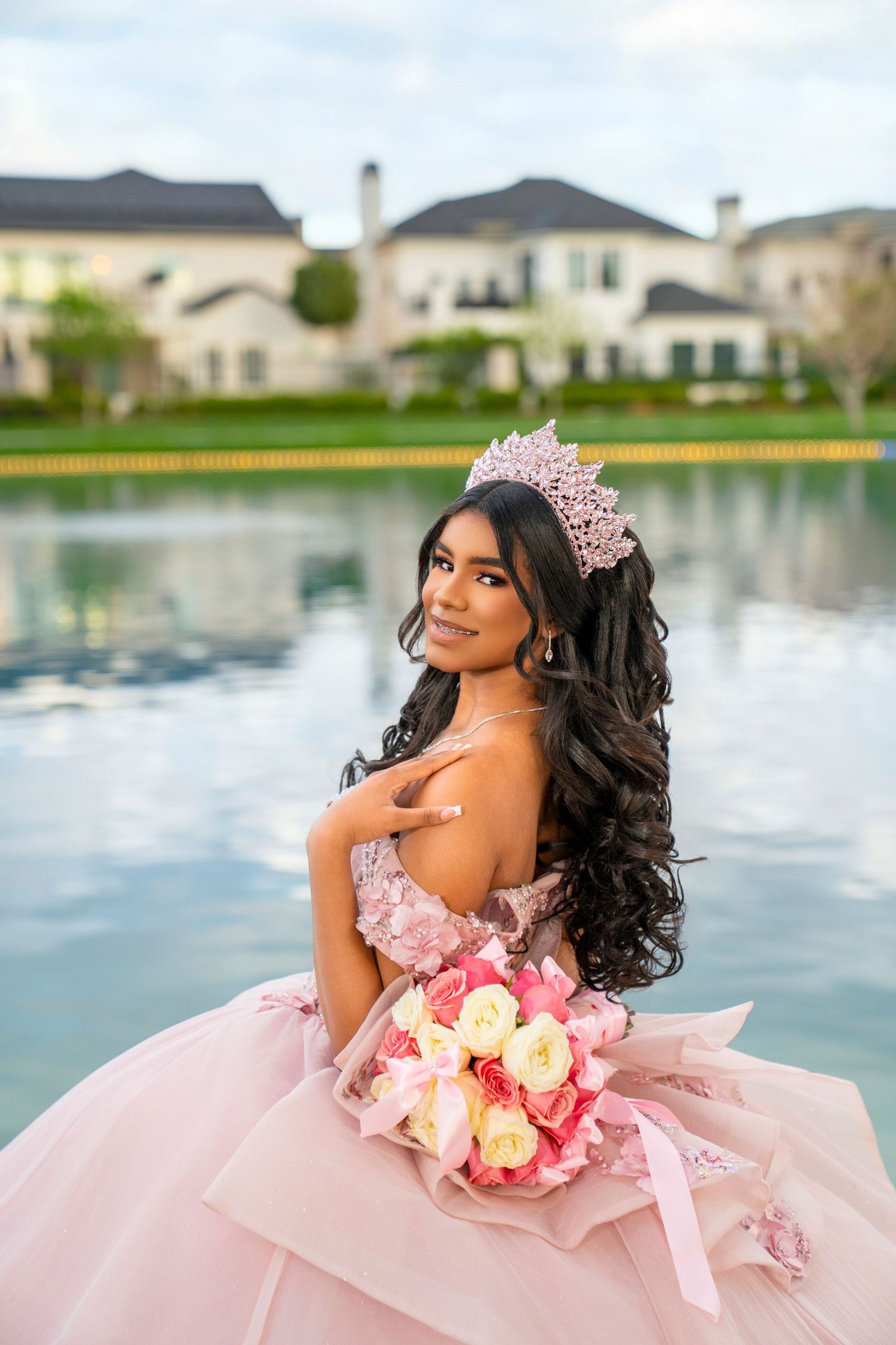 Young woman in pink quinceañera gown and floral crown holds bouquet by a reflective pond with elegant homes backdrop.