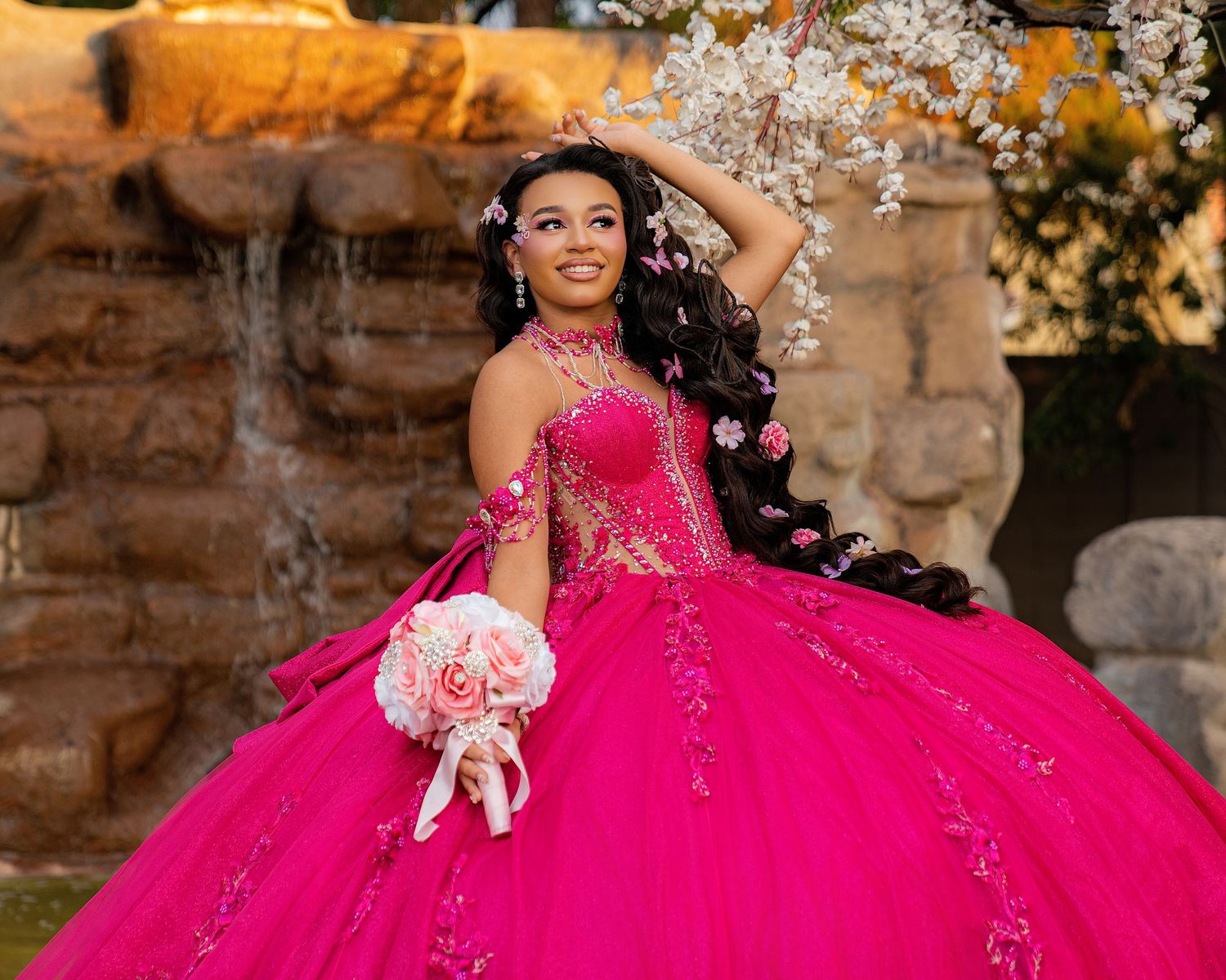 Young woman in a fuchsia ball gown quinceanera dress with floral details, holding a bouquet near a fountain.