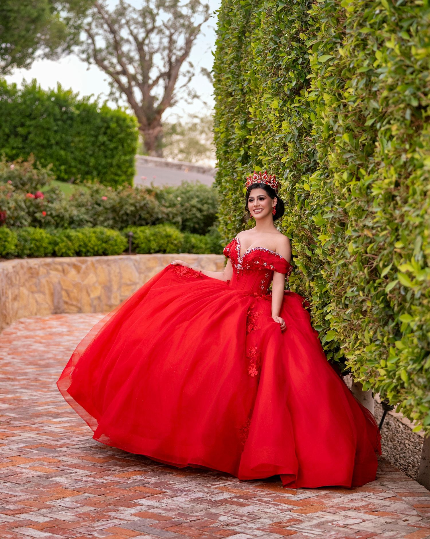 Young woman in a stunning red off-shoulder ball gown quinceañera dress poses in a lush garden setting.